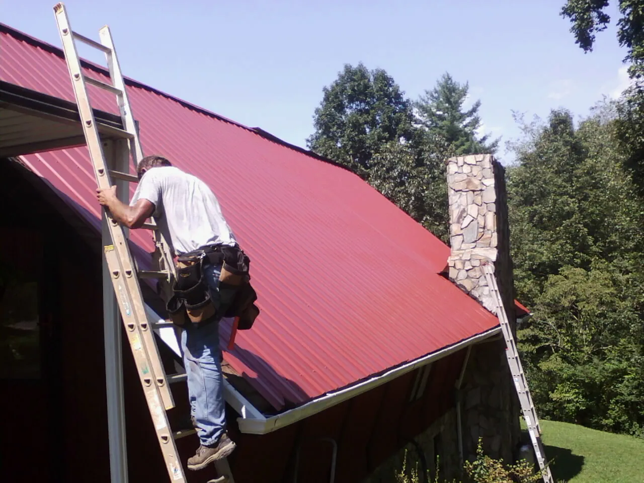 Roofer on a ladder beside a red metal roof and stone chimney — Soffit & Fascia Repair by Roofing Brothers in Lincoln Park